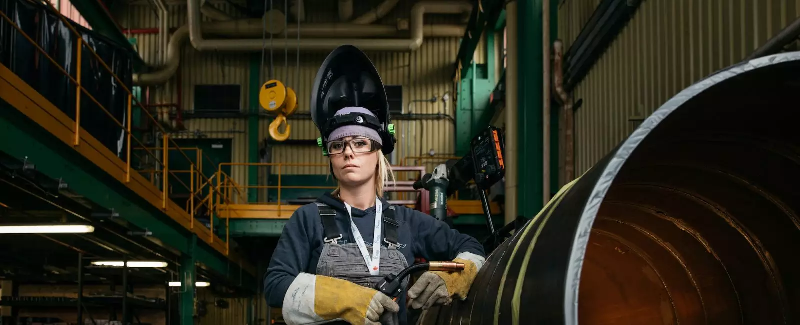 A woman in welding gear standing beside a large metal pipe